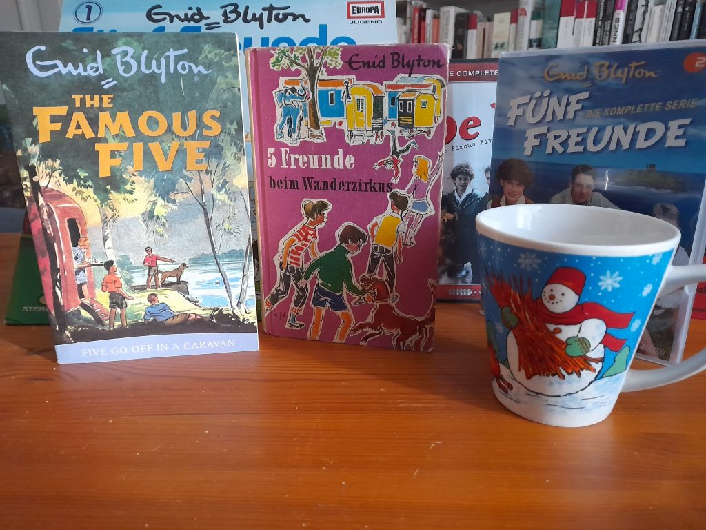 Das Bild zeigt zwei Bücher. Eins ist die englische Ausgabe "Five go off in a Caravan", das andere ist die deutsche Ausgabe "5 Freunde beim Wanderzirkus". 